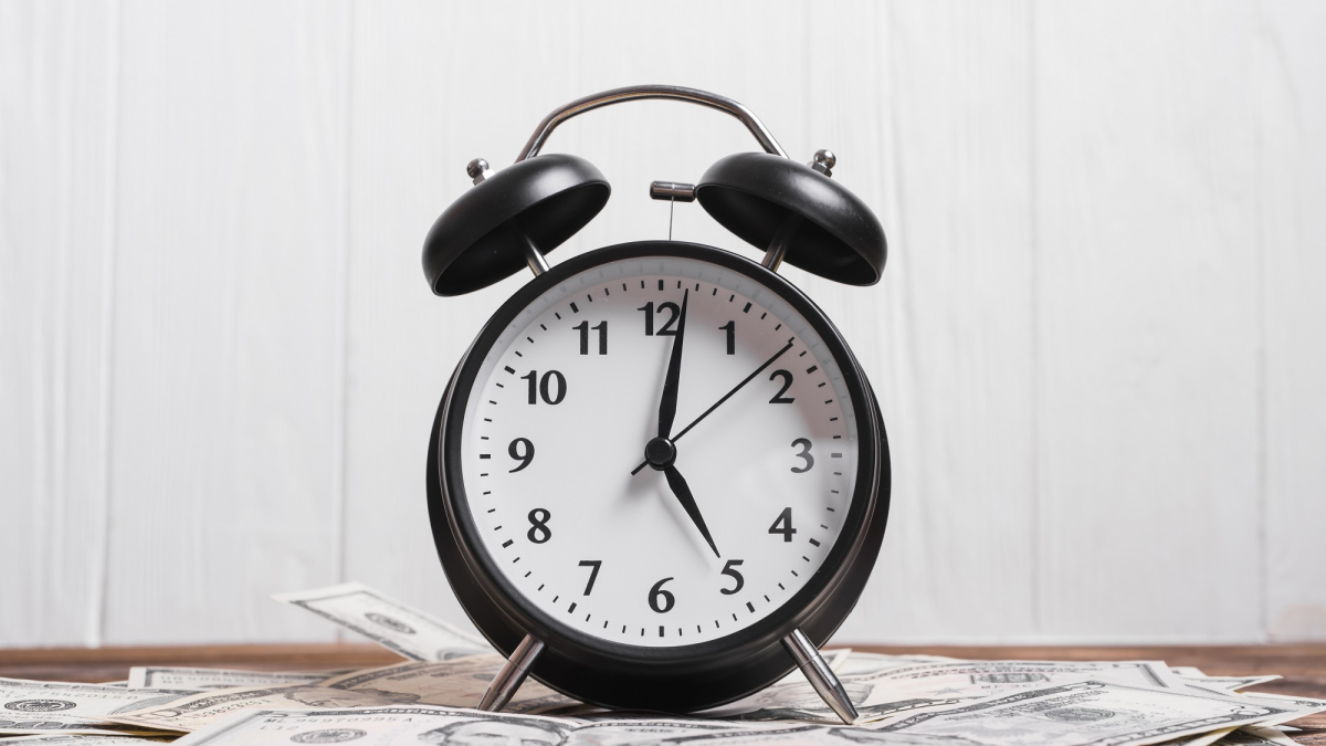 alarm-clock-us-dollar-currency-notes-wooden-desk-against-white-wall